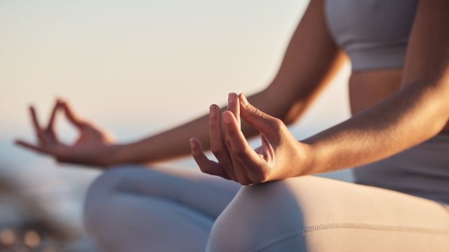 A person in yoga pose outdoors, hands in a mudra/gentle meditation gesture, seated in a calm, sunlit setting by the water.