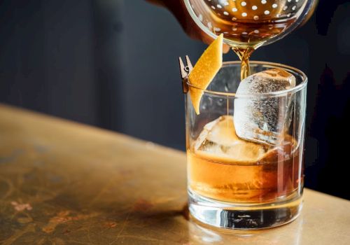 A glass with whisky over ice, garnished with an orange twist, as a golden stream pours from a bottle above.