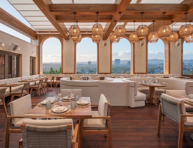 A bright, airy restaurant with wooden beams, warm hanging pendant lights, large windows showing city views, and cozy white and beige seating.
