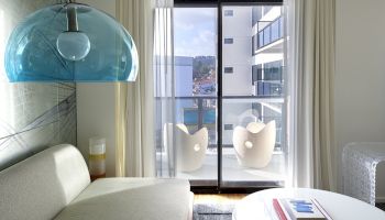 A modern living room with a white sofa, a blue glass pendant light, a round white coffee table, and floor-to-ceiling glass doors opening to a balcony with white chairs, overlooking buildings.