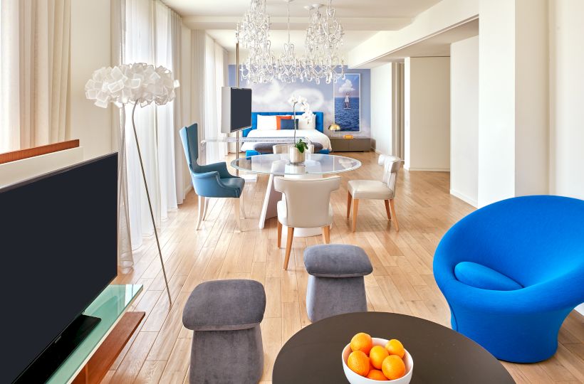 A bright, modern living room with a round table and bowl of oranges in the foreground, blue chair, gray stools, and a chandelier overhead.