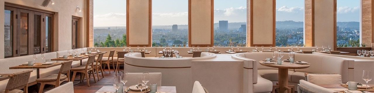A stylish rooftop dining area with wooden beams, wicker chairs, soft beige seating, round pendant lights, and city skyline views in the distance.