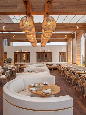 A stylish, sunlit restaurant with curved white booths, wooden tables, and wicker pendant lights under a airy wooden pergola, inviting dining.