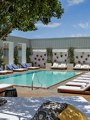 Hotel pool area with a rectangular pool, lounge chairs along both sides, shaded cabanas, and a few seating cushions on a deck under clear blue skies.