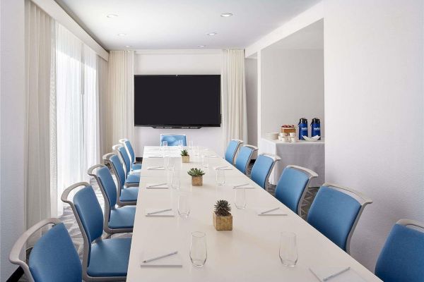 A modern conference room with a long white table, blue chairs, a wall-mounted TV, and natural light from large windows, set for a meeting.