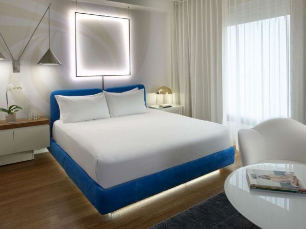 A modern hotel room with a blue-upholstered bed, white linens, soft lighting, wall-lit square art, sheer curtains, and a small seating table.