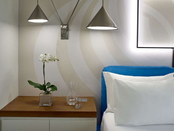 A neatly made hotel bed beside a wooden nightstand with an orchid, two glass cups, and modern pendant lights overhead.