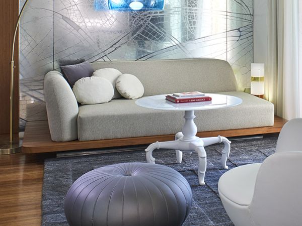 A modern living room with a light gray sofa, round pouf, white coffee table, futuristic wall art, and sculptural lighting in a bright, airy space.
