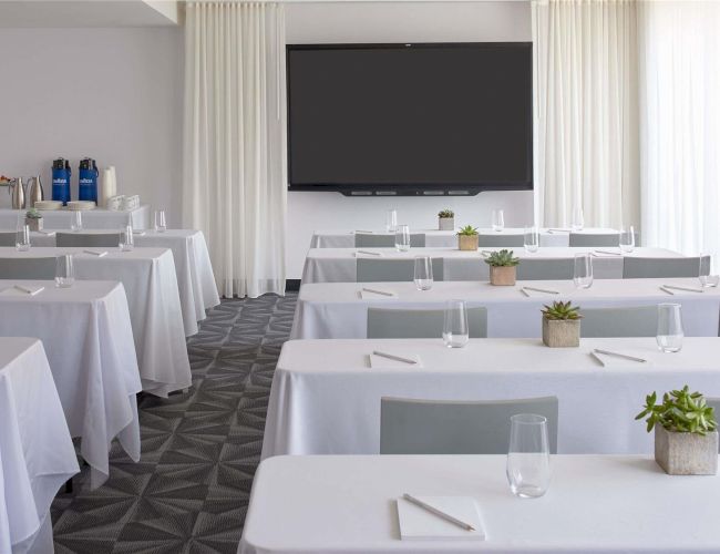 A modern conference room set for a formal event with white-clothed tables, small plants, and a large screen at the front ending the sentence.