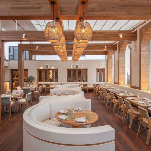 A stylish, airy restaurant with warm wood tones, curved white seating, and long communal tables under woven pendant lights, creating a cozy outdoor‑in vibe.