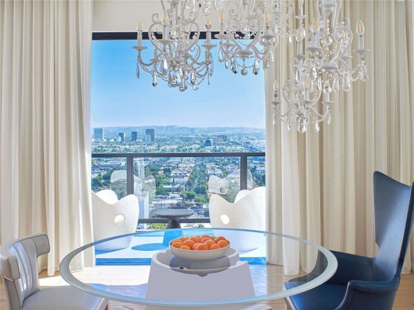 Luxurious dining nook with a glass round table, chandelier, plush chairs, sheer curtains, and a panoramic city view from a bright high-rise balcony.