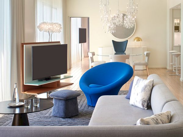 A modern living room with a gray sofa, blue accent chair, flat-screen TV, glass coffee table, and a bright dining area in the background.