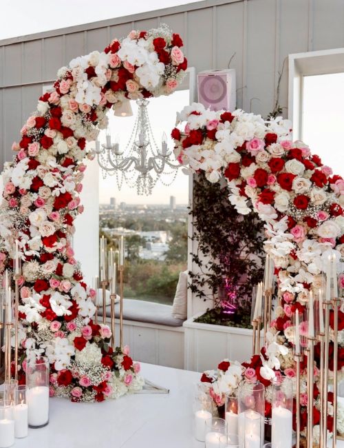 A floral heart arch made of red, white, and pink blooms with hanging crystals and candles along a windowsill wedding backdrop.
