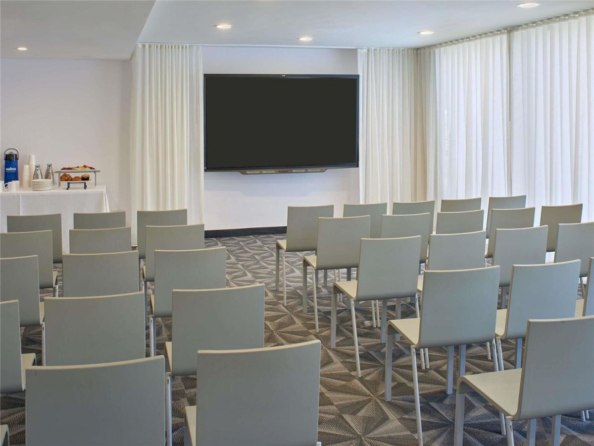 A modern conference room with a large screen, white curtains, and rows of light gray chairs facing the presentation area.