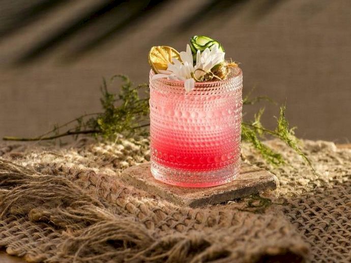 A pink cocktail in a textured glass with a lime wedge and mint leaves, on a woven burlap mat outdoors.