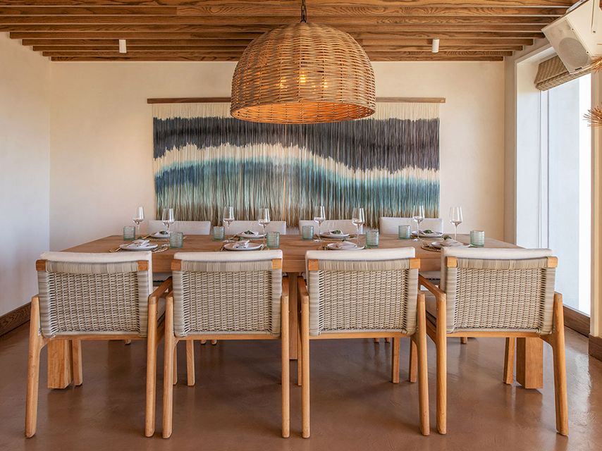 A bright dining room with a wooden table set for eight, wicker chairs, a woven pendant light, wall art, and natural light from large windows.