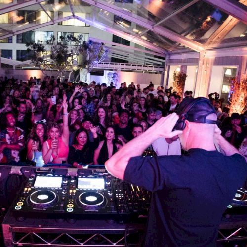 A DJ spins at a crowded indoor party with a large cheering audience under colorful lighting, mixing tracks on professional gear.