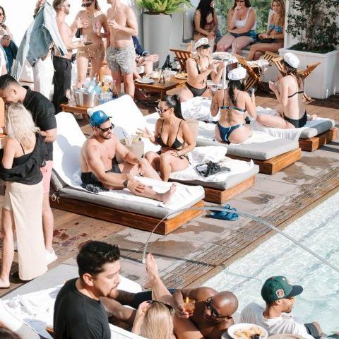 A crowded pool party with many people lounging on sunbeds, chatting, and relaxing near and around the poolside.