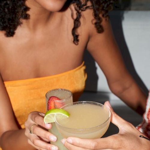 Two friends toasting cocktails with lime wedges; one in an orange dress shows a close-up of hands and drinks.