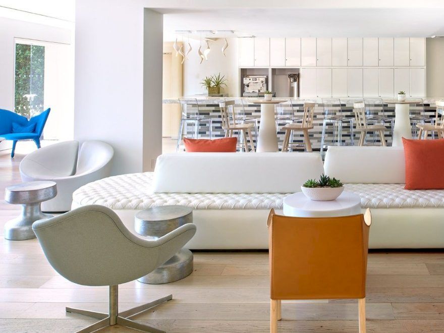A modern, airy living room with white furniture, a patterned sofa, neutral chairs, orange throw pillows, and a bright kitchen/bar area in the background.