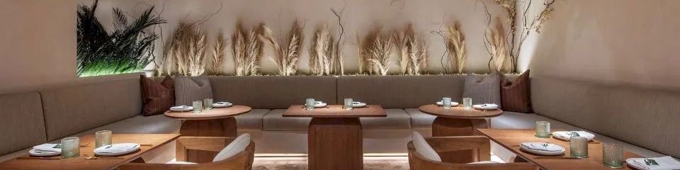A modern, minimalist restaurant patio with wooden tables, white chairs, cushioned bench seating, and decorative tall grasses along the back wall, under a wooden beam ceiling.