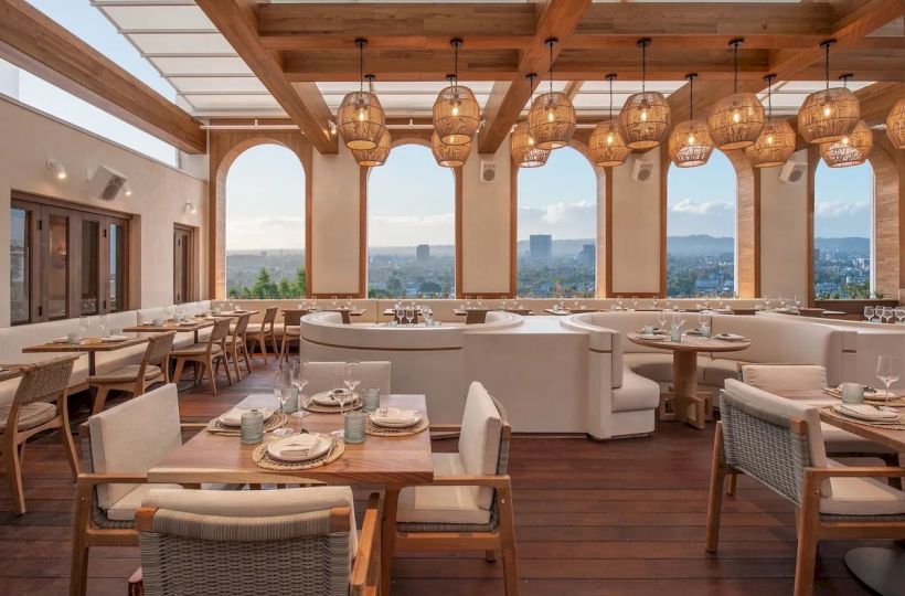A stylish rooftop restaurant with wooden beams, warm hanging lanterns, cushioned seating, and panoramic city views; chic casual dining space.