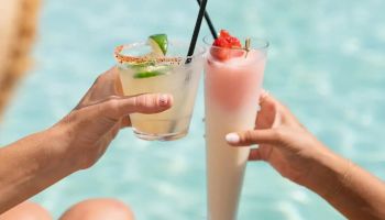Two people clink cocktails by a pool: one lime-tinted drink with a lime wedge, the other a tall pink beverage with a strawberry, both with black straws.