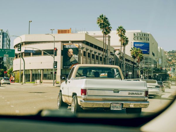 A white vintage pickup driving on a sunny city street, viewed from inside another car, with palm trees and storefronts in the background.
