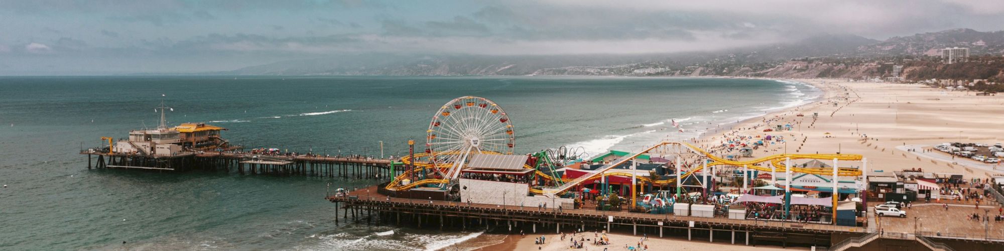 A crowded sunny beach with a long pier, colorful rides and a Ferris wheel at the end, waves washing onto the sandy shore, vacation vibes.