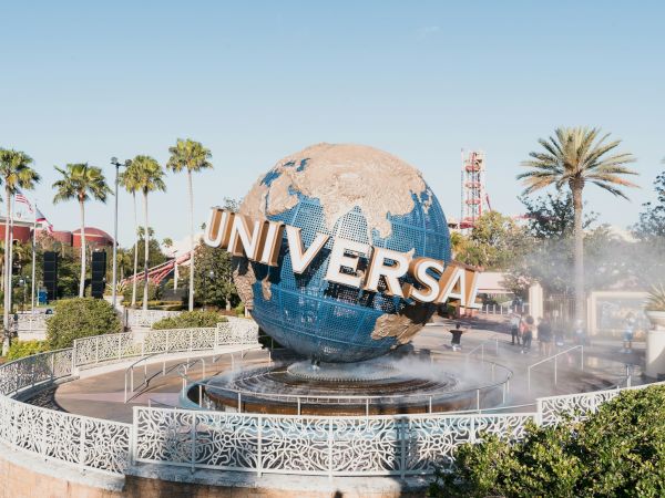 A globe sculpture reading &ldquo;UNIVERSAL&rdquo; at a fountain with palm trees around it, in a sunny theme park setting.