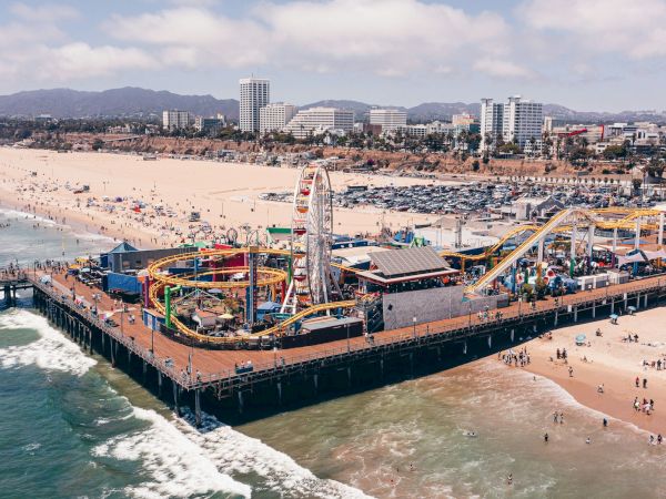 A sunny beach with a bustling pier and amusement rides, including a ferris wheel, roller coasters, and crowds enjoying the ocean breeze.