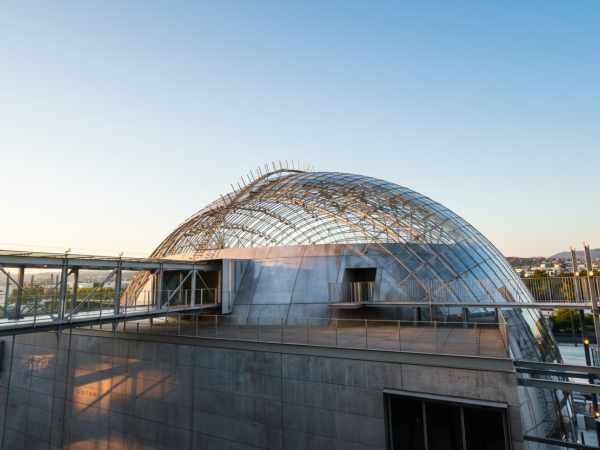 A glass-domed arena or observatory atop a concrete building, with a curved lattice roof, sky-blue backdrop, and a bridge-like walkway surrounding it.