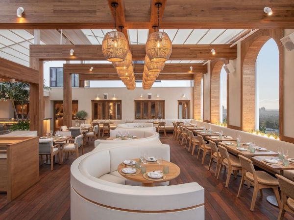 A stylish, airy restaurant with wooden beams, warm lighting, curved white seating, and long wooden tables set for dining, overlooking a bright outdoor view.