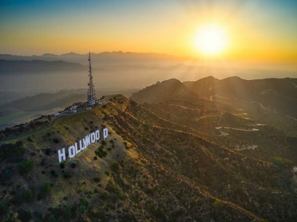 A hillside overlooking Los Angeles at sunrise with the famous HOLLYWOOD sign and a tall antenna perched on the ridge, bathed in golden light.