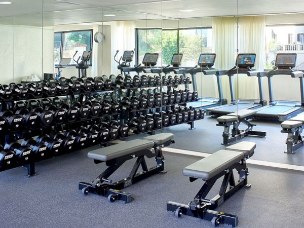 A spacious gym with a row of treadmills and a wall of dumbbells, plus adjustable benches and mirrors reflecting the workout space.