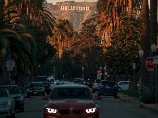 A sunset-lit street near the Hollywood Hills, cars on the road, palm trees lining the avenue, and the Hollywood sign visible in the distance.