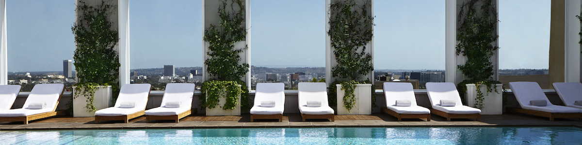 A rooftop pool deck with white lounge chairs, potted trees, and large windows reflecting the sunny sky, overlooking a distant cityscape.
