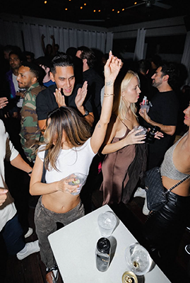 People dancing at a crowded party, raising hands, drinks on a table, energetic nightlife scene.