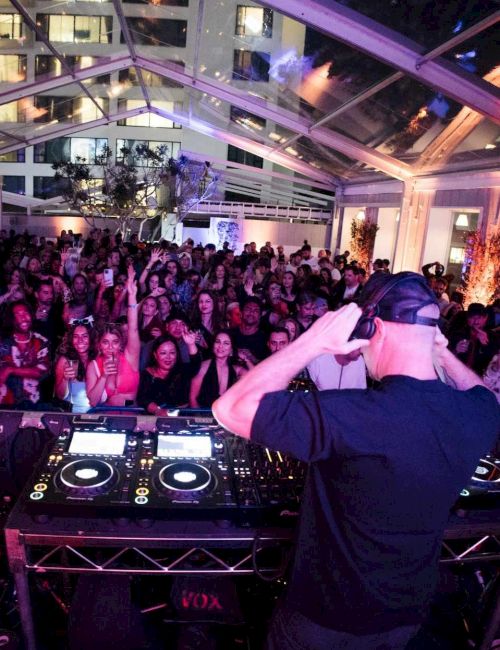 A DJ performs at a crowded indoor party, wearing a cap, blending tracks on turntables while a packed audience cheers and watches the stage.