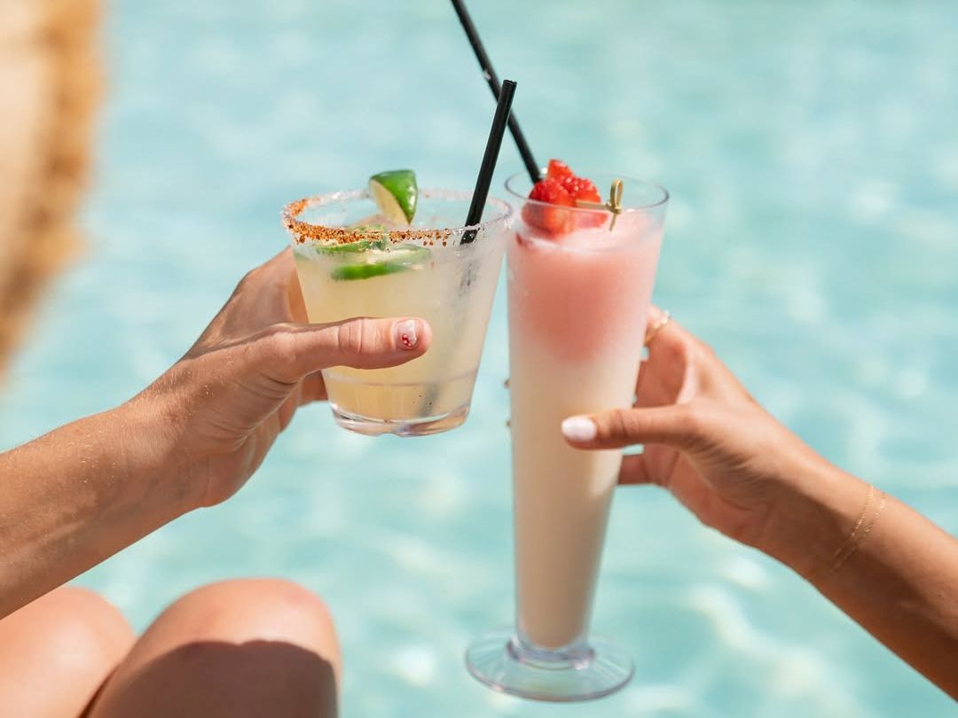 Two people clink tropical cocktails by a pool, hands holding fancy drinks with straws, lime and strawberry garnishes, sunny vibes.