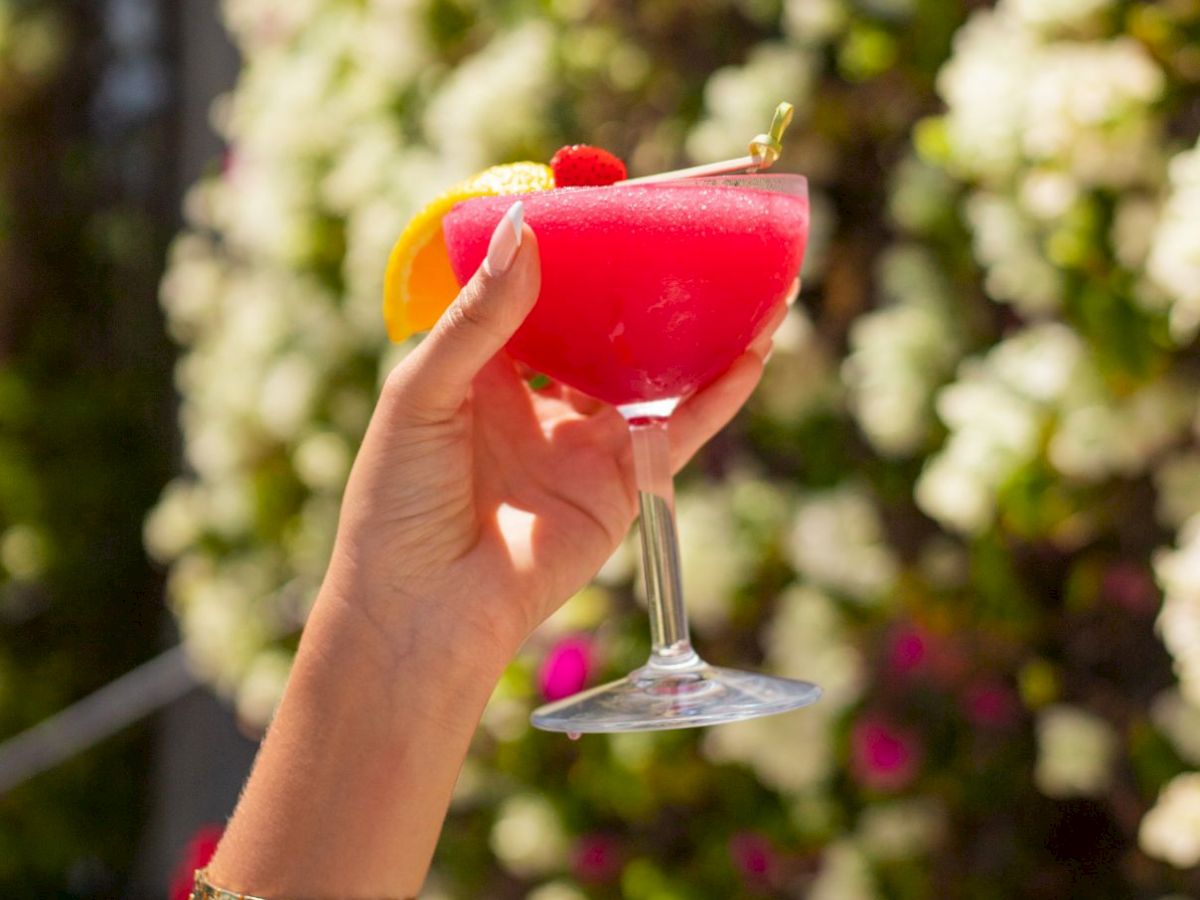 A hand holds a pink cocktail with an orange slice, set against a sunny garden background.