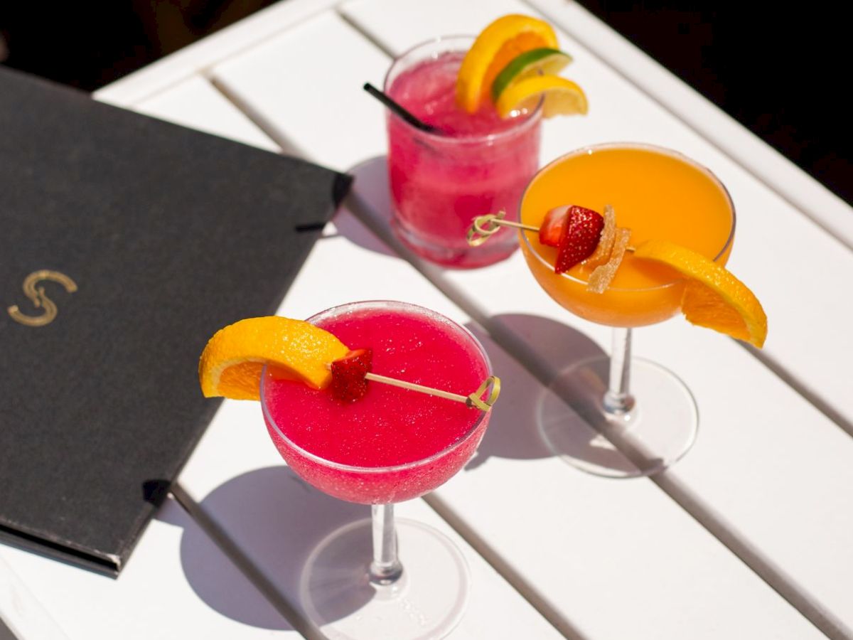 Three colorful cocktails with orange garnish sit on a white table beside a black menu; sunny outdoor vibe, bright and inviting.