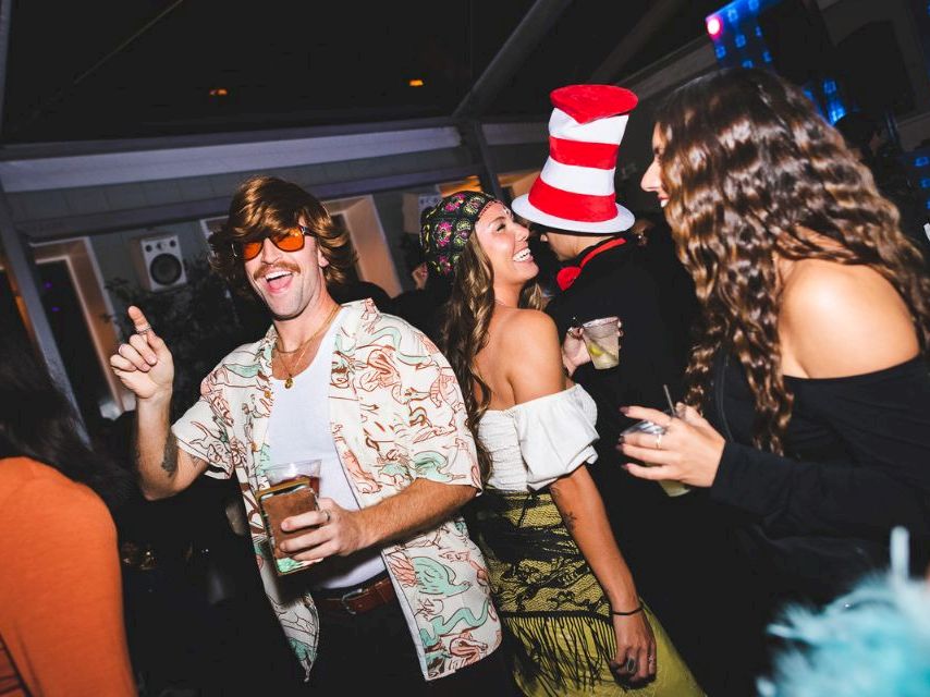 A lively party scene with stylishly dressed people dancing and chatting, some wearing quirky hats, enjoying drinks in a crowded club vibe.