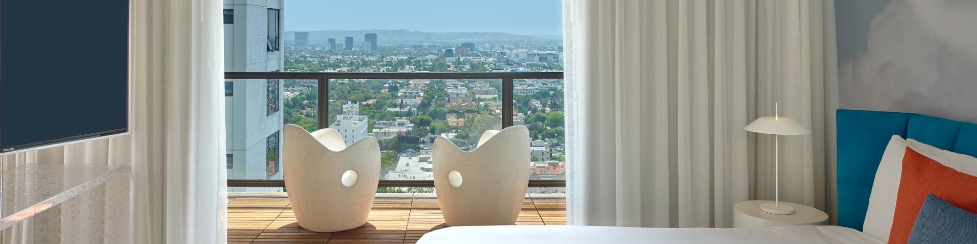 A bright hotel room with a large window view, a bed with white linens and blue/orange pillows, a TV, a bench, and a balcony with round chairs.