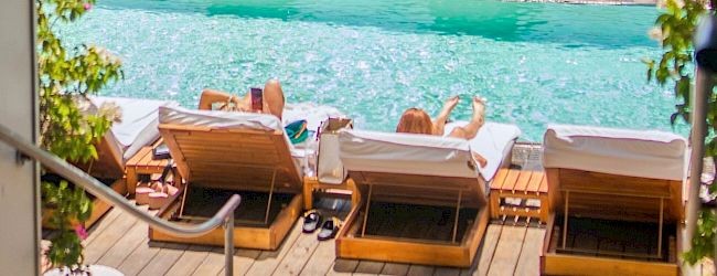 A luxurious poolside scene with lounge chairs, a sparkling turquoise pool, and leafy planters against a mural-covered wall.