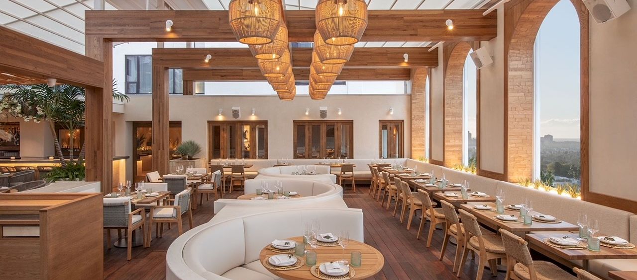 A stylish, bright restaurant with warm wood tones, curved white booth seating, long rows of tables, hanging woven light fixtures, and large windows.