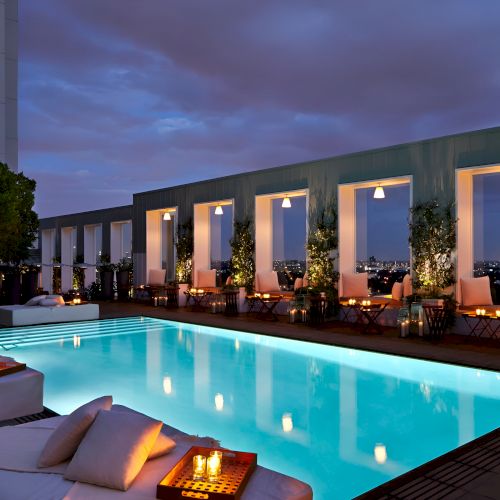 A rooftop pool with lounge beds, warm lighting, and seating along the water, overlooking a city skyline at dusk.