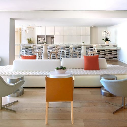 A modern, open-concept living area with a large white sofa, light wood floors, and minimalist chairs; a bright kitchen bar in the background.