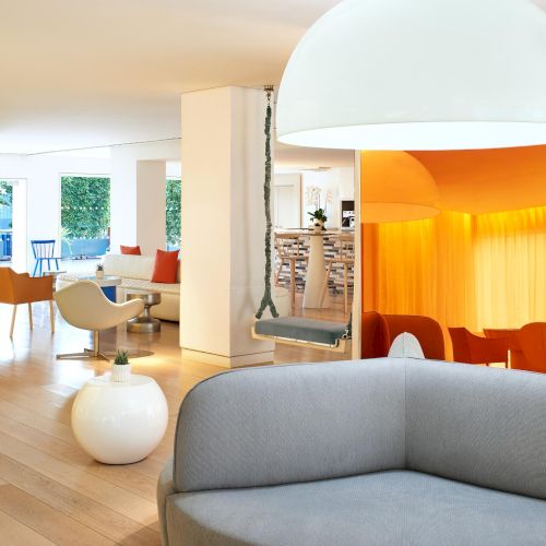 A bright, modern lounge with colorful chairs, a curved gray sofa in foreground, large white pendants, wooden floors, and a warm orange seating nook.