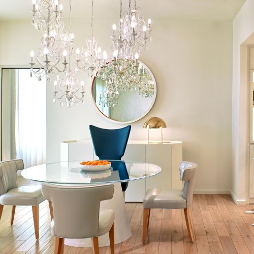 A bright, modern dining area with a round glass table, beige chairs, chandelier lighting, and a mirrored wall accent. Top it at 140 characters, always ending the sentence.
